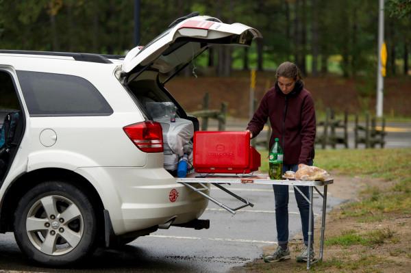 Claire from @carnet_declaireurs setting up a camping stove and preparing a meal at the back of a white SUV during a camping adventure in Canada. The vehicle's trunk is open, revealing camping gear and supplies, capturing the essence of mobile travel and outdoor cooking in a parking lot in Canada.