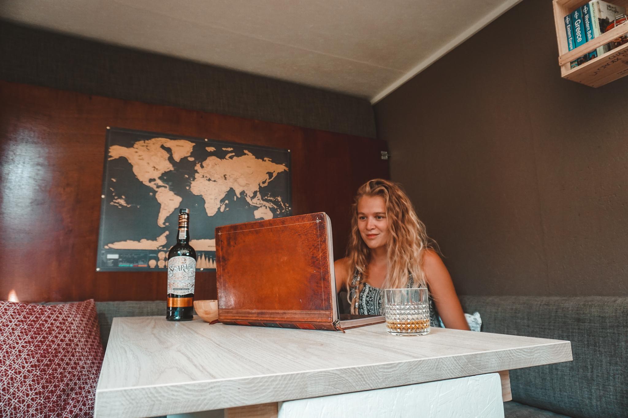 Young woman working on a laptop with a vintage book-style cover, seated at a table inside a tiny home. The table features a bottle of Scotch and a glass of whiskey. In the background, a world map hangs on the wall, adding a cozy, traveler's vibe to the compact, wood-paneled interior.