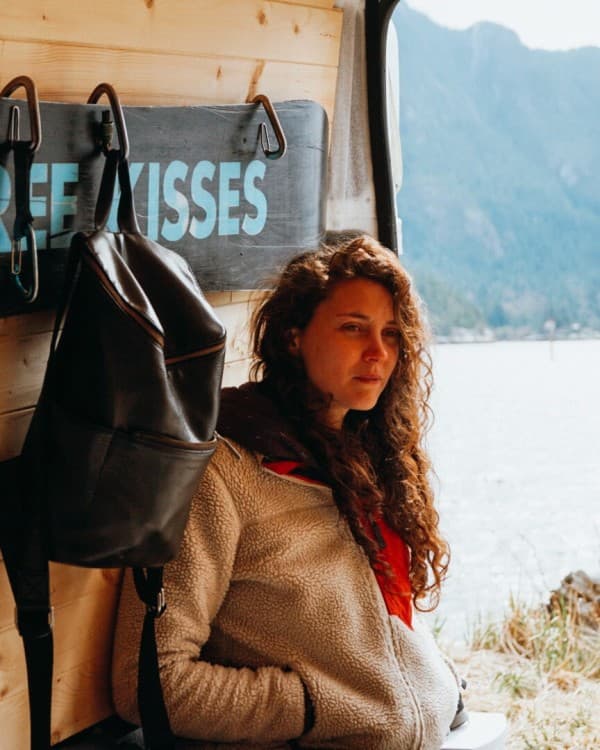 A woman, Georgia Broderick, in a casual pose inside her travel van, with the back doors open to a scenic lake and mountain landscape. A unique 'Free Kisses' snowboard is displayed beside her, with a stylish black backpack hanging nearby, blending adventure lifestyle and nature in one compelling image.