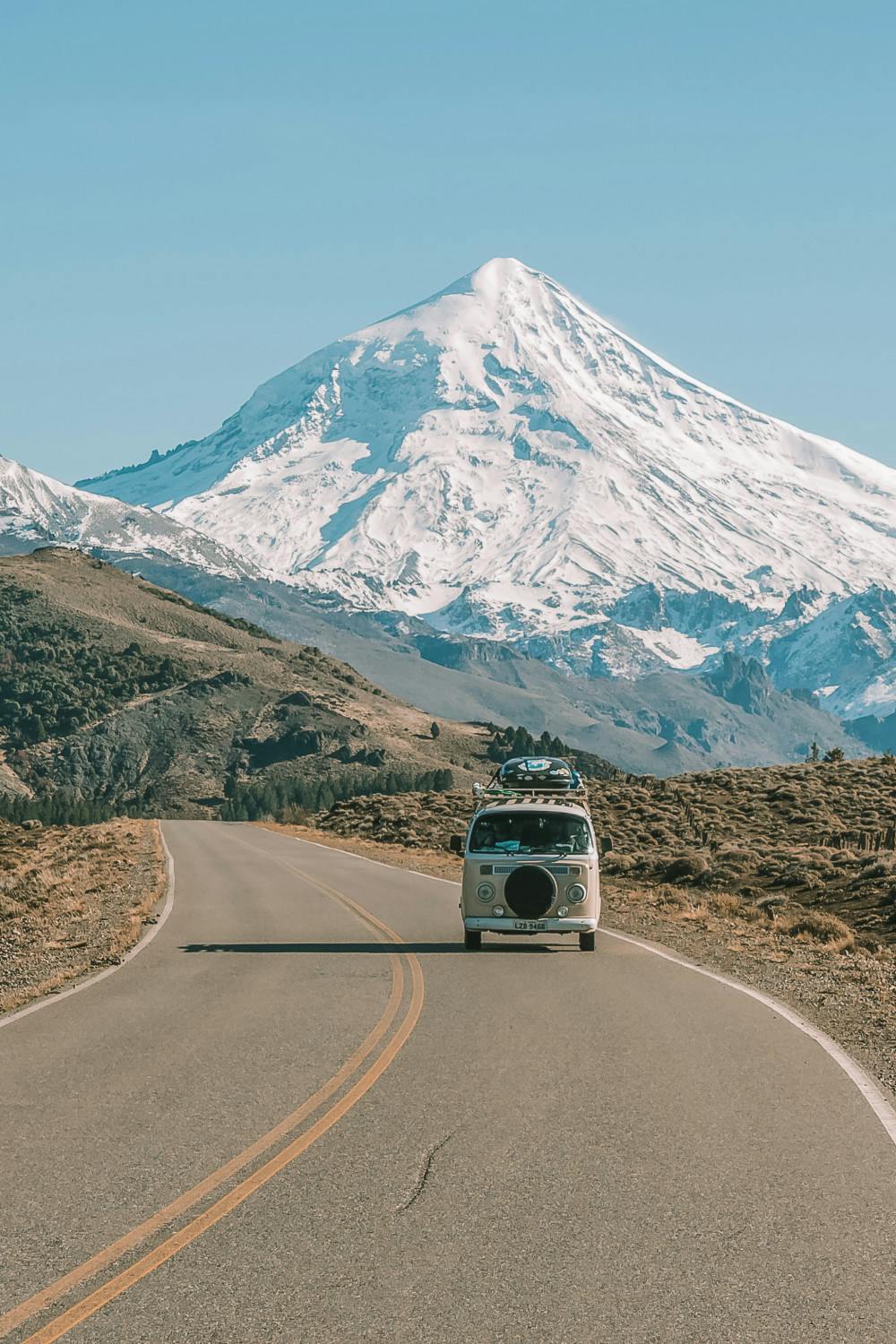 A classic Volkswagen van with luggage on the roof rack drives on a scenic highway away from a majestic, snow-capped mountain. The road curves gently in the foreground, framed by rugged terrain and clear blue skies, invoking a sense of adventure and travel in the great outdoors.