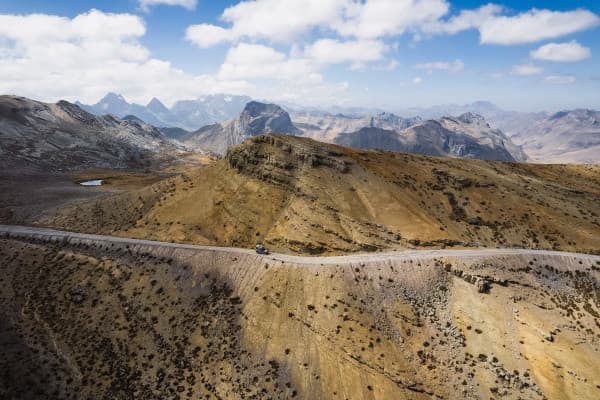 A rugged Puch 230GE camper driving along a narrow mountain road through remote, rocky terrain in the Andes, South America, with expansive mountainous landscapes in the background.