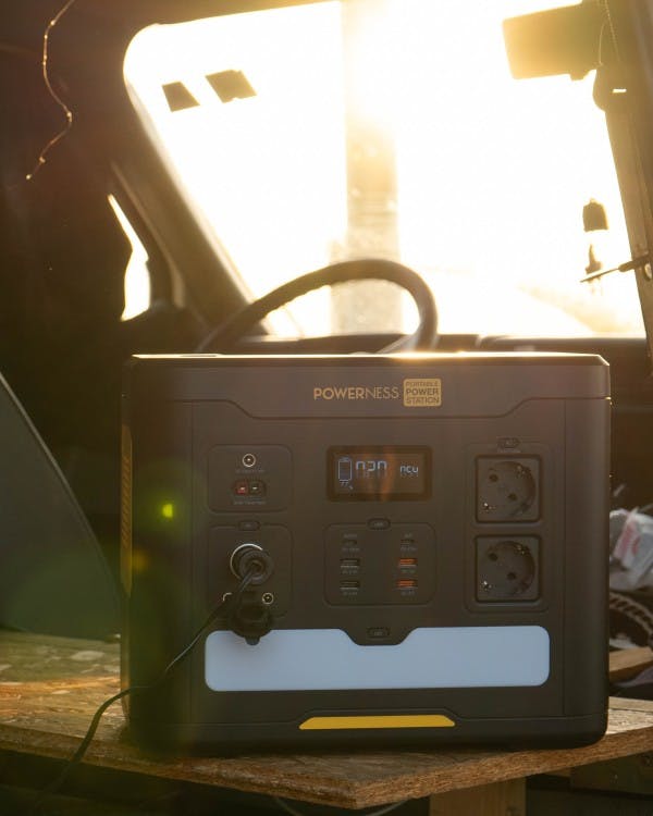 This image shows a portable power station inside a van, with a warm sunset or sunrise visible through the windshield. The power station is placed on a wooden table or platform, indicating a possible van conversion for travel or camping. The power station is equipped with various outlets and ports, ready to provide off-grid electrical power. The sunlight glinting through the window adds a feeling of adventure and self-sufficiency.