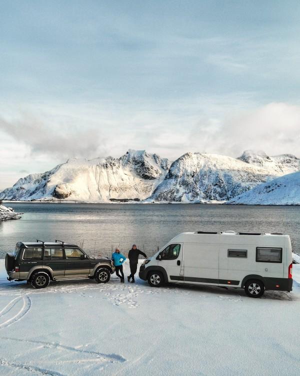 Winter adventure in Northern Norway with a campervan and SUV parked by a serene snowy lake surrounded by stunning snow-capped mountains. Capturing the essence of travel and exploration in the Arctic by @travelvan.