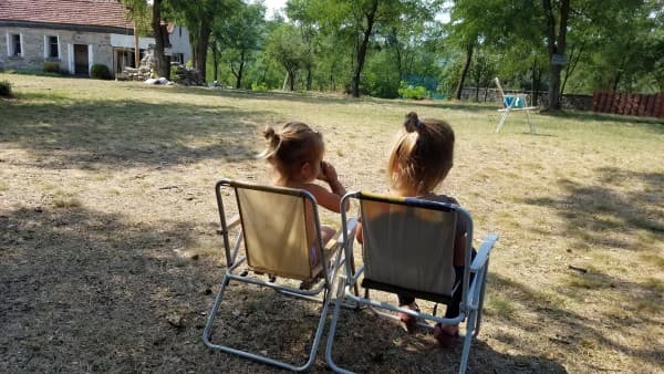 Two young girls, Stella and Zara, sit side by side in small camping chairs in a sunny, rural yard in Bulgaria. They are enjoying a quiet moment together, surrounded by trees and a rustic stone house in the background. The scene captures a peaceful and simple moment of childhood during their family's van life adventures.