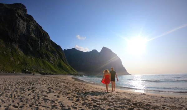 A couple, @travelvan, enjoying a serene walk on a sandy beach in Norway at sunset. The image captures the stunning contrast of the beach, the towering green mountains, and the calm ocean, highlighting the beauty of Norway's coastline.