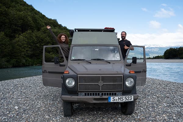 Sarah and Mats standing in the open doors of their Puch 230GE 4x4 offroad camper, parked on a rocky riverbank in South America, with a backdrop of forested mountains and a clear river.