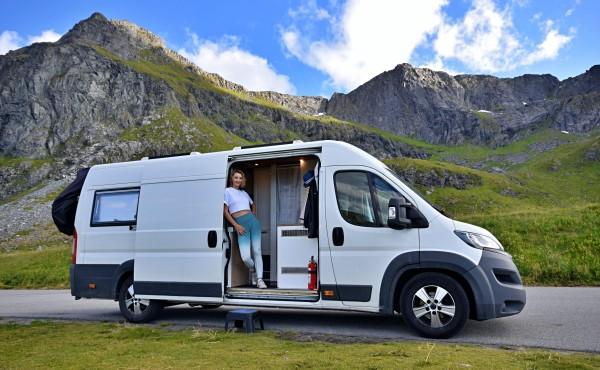 A young woman, Mary from @travelvan, standing in the doorway of a self-converted campervan set against the backdrop of lush green mountains in Norway. The image captures the essence of freedom and adventure on the road.