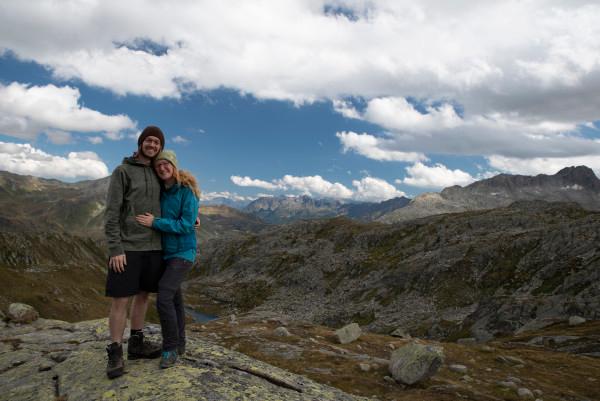 Christina and Ralf, known as @miles_together, are standing together and smiling on a rocky outcrop in the Gotthard region of Switzerland. They are surrounded by a stunning mountainous landscape under a partly cloudy sky. The image captures their adventurous spirit and love for exploring nature.