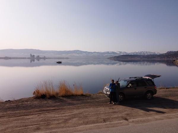 David and Claire from @carnet_declaireurs standing beside their SUV with the trunk open, overlooking the serene Okanagan Lake in Canada. The calm lake reflects the distant mountains and clear sky, capturing the peaceful and scenic essence of their vanlife adventure. The image showcases the beauty of Okanagan Lake and the couple's love for travel and exploration.