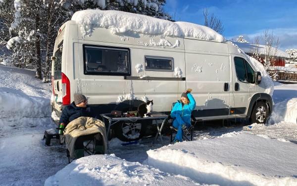 Mary, Peter and their cat Stefcia enjoying a snowy winter day outside their converted campervan in Norway. The van is covered in snow, highlighting the adventurous spirit of winter van life in the Arctic.