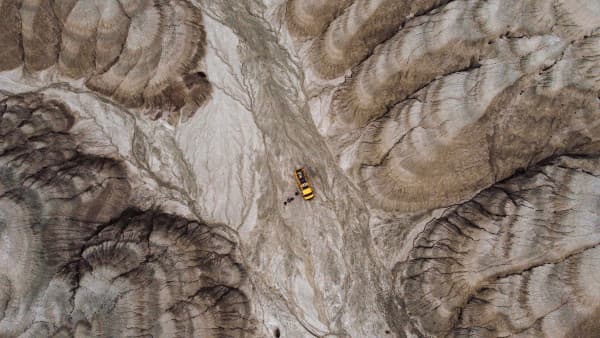 Aerial view of a yellow Mercedes Vario 816D camper van from @our_orbit_vanlife parked in a dramatic, rugged landscape with eroded rock formations. The van is situated in the center of the image, highlighting the stark contrast between the bright vehicle and the earthy tones of the surrounding terrain, showcasing the adventurous spirit of vanlife in remote, natural settings.