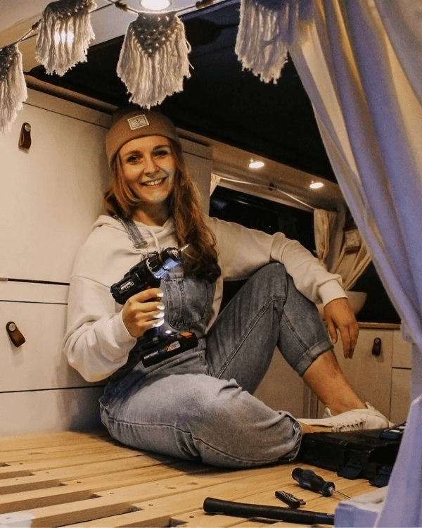 A woman (@itsme.jenni) sitting inside her self-built camper van, holding a power drill. She is smiling and appears proud of her work. The van interior includes wooden furniture and cozy decor, reflecting her journey of transforming a VW T5 into a mobile home.