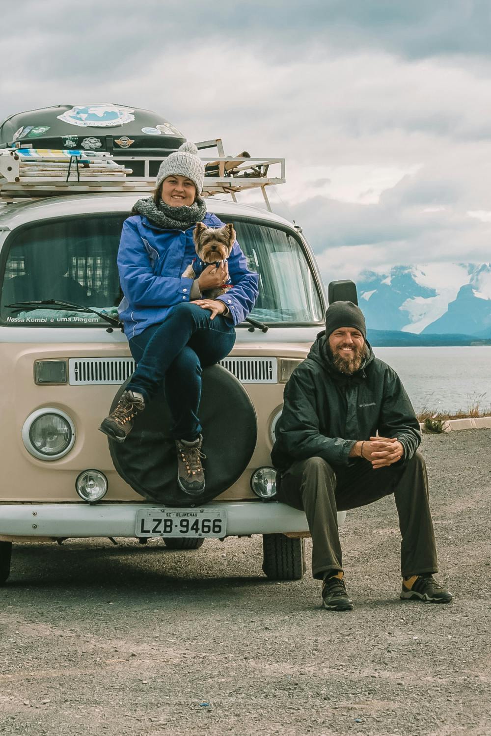 A smiling woman wearing a blue jacket and a beanie sits on the front tire of a vintage cream-colored Volkswagen van, holding a small Yorkshire Terrier. A man with a beard, also smiling, sits on the ground leaning against the van, wearing a black beanie and jacket. They seem to be enjoying a chilly day with a scenic backdrop of mountains and a cloudy sky. The van has a roof rack with assorted gear, suggesting an adventurous road trip.