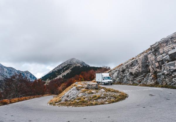 White box truck driving on a winding mountain road with rocky terrain and autumn-colored trees, under a cloudy sky. The road curves around a hill with a prominent peak in the background, showcasing a dramatic and scenic mountainous landscape.