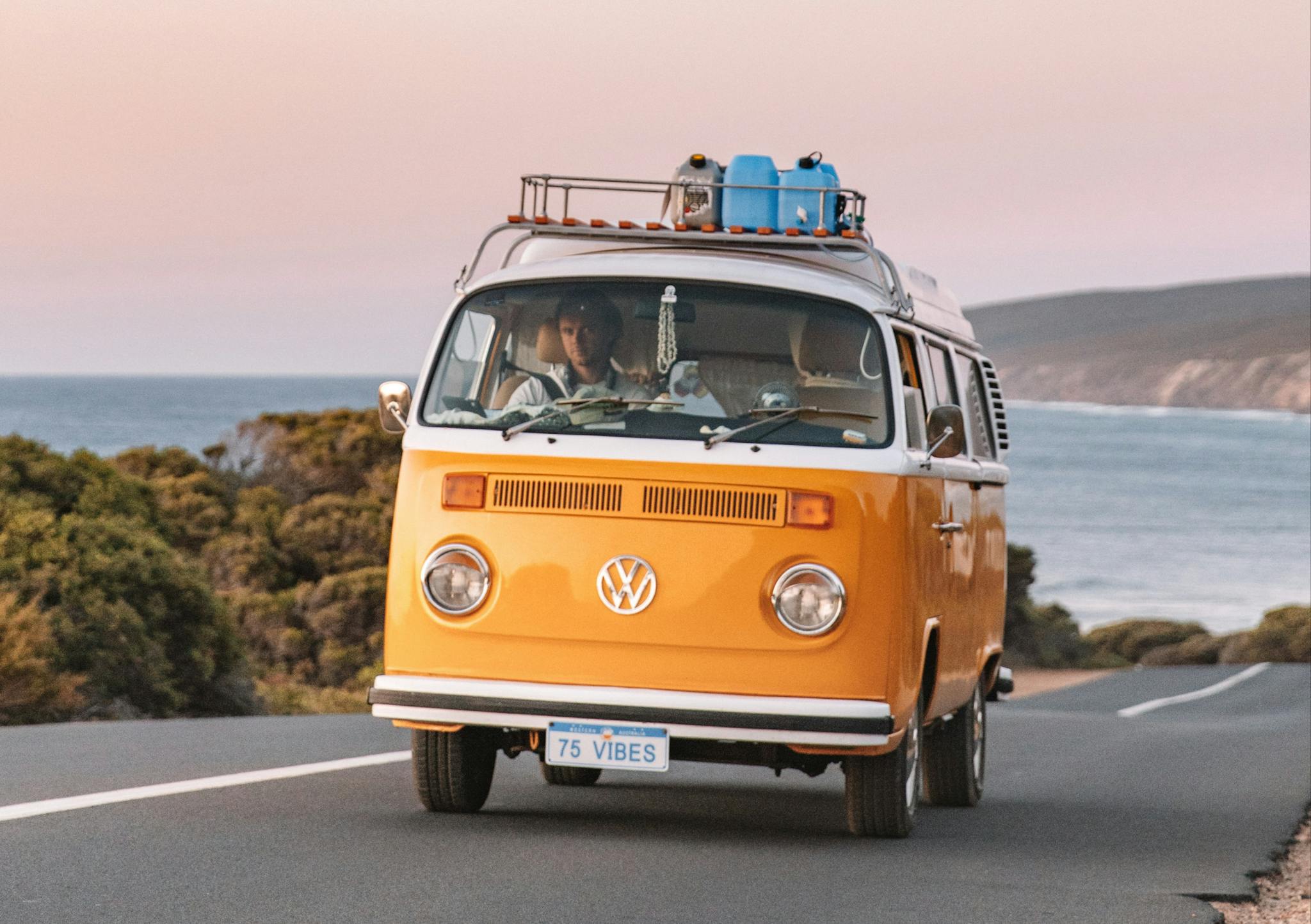 A yellow Volkswagen van with the license plate '75 VIBES' drives along a coastal road. Surfboards are visible on the roof rack, hinting at an adventurous beach outing. Inside, a person is focused on the road ahead, while the backdrop features a calm sea and a clear sky at twilight, conveying a sense of freedom and the spirit of a road trip along the picturesque coastline. @75vibes_ 09/2020 - Vanlifezone (Issue 1)