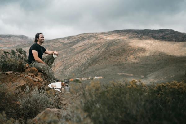 A man sits on a rocky outcrop in the mountains, enjoying a break during his outdoor adventure. He is dressed in casual hiking attire with a black t-shirt and khaki pants, holding a cup of what appears to be a hot beverage. The landscape behind him features rolling hills and a cloudy sky, creating a serene and picturesque scene. Camping equipment, including a portable stove, can be seen beside him, indicating a well-prepared hiking or camping trip. This image captures the essence of outdoor exploration and the tranquility of nature.