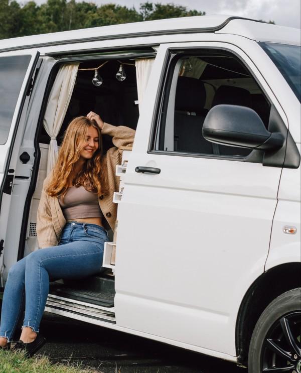 A woman (@itsme.jenni) sitting in the doorway of her self-built camper van, enjoying the outdoors. She is smiling and relaxed, with the van's interior partially visible behind her. 