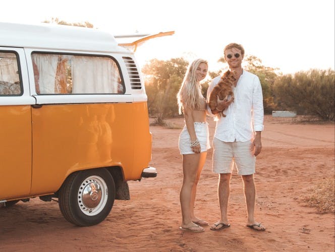 A young couple stands smiling beside a classic half white, half yellow van on a sandy terrain in Australia, bathed in the warm light of the setting sun. The woman, dressed in white shorts and a top, holds a small, fluffy brown dog in her arms. The man, wearing sunglasses, a white shirt, and beige shorts, stands beside her with a relaxed posture. Trees in the soft-focus background suggest a remote, tranquil location. @75vibes_ 09/2020 Vanlifezone (Issue 1)