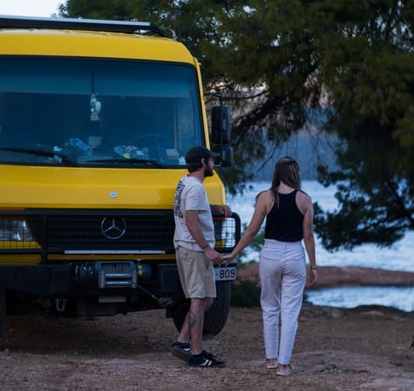 A couple, @our_orbit_vanlife, stands hand in hand beside their yellow Mercedes Vario 816D camper van parked in a scenic outdoor setting with trees and a body of water in the background. 