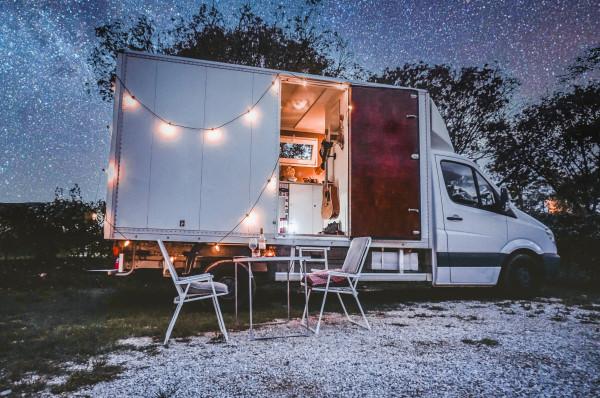 Cozy mobile tiny home converted from a white cargo van parked under a starry night sky, featuring an open back with string lights, a comfortable interior setup visible through the back doors, and outdoor seating area with two white chairs and a small table on a gravel surface.