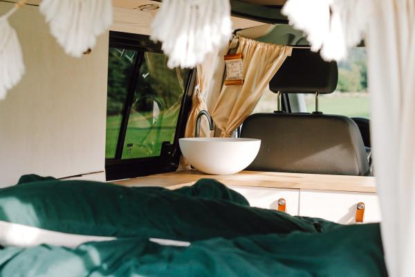 The interior of a self-built VW T5 camper van, featuring a neatly arranged sink, wooden countertop, and cozy green bedding. The van, transformed by Jenni (@itsme.jenni), showcases her DIY skills and attention to detail.