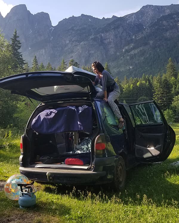 A woman is setting up a makeshift sleeping area in the back of a Renault Espace microcamper parked in a scenic mountainous area in Bulgaria. The van's trunk is open, revealing bedding and travel gear inside, with lush greenery and towering mountains in the background. A portable gas stove and a toy ball are placed on the grass next to the van.