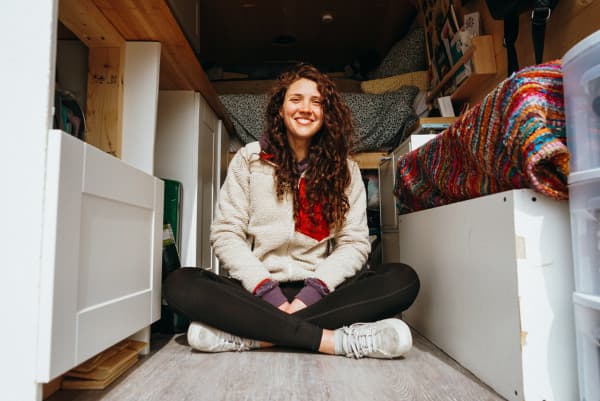 Georgia Broderick sits cross-legged and smiling inside her van, showcasing the efficient use of space with visible storage solutions. The van's cozy interior includes a colorful knitted blanket and an assortment of personal items, capturing a moment of joy in the compact living area of her home on wheels, ideal for those interested in van life and travel.