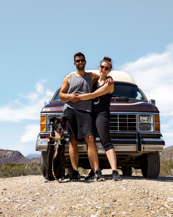 A happy couple with their dog standing in front of a classic 1985 Dodge B250 van on a dirt road in Texas, with clear blue skies and desert landscape in the background.