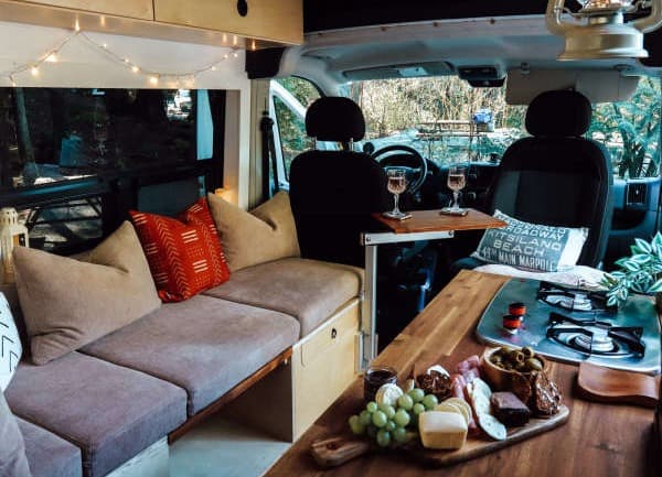 Cozy interior of Emily Jay's RAM Promaster, featuring a comfortable seating area with cushions, string lights, and a small dining setup. The wooden countertop has a charcuterie board with various snacks, and a portable stove. The inviting space showcases the practical and charming aspects of vanlife, set against a backdrop of the outdoors in Canada.