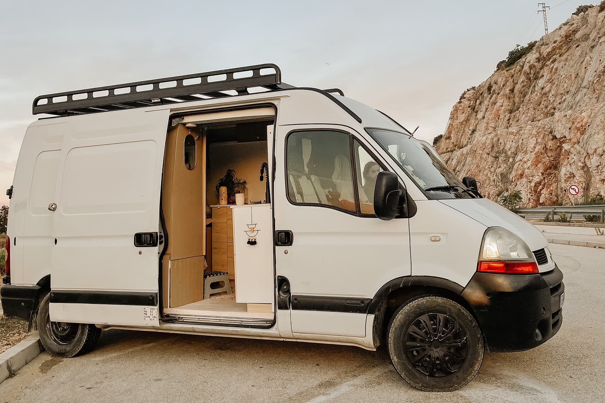 The converted Renault Master by @irisandmax van parked with its side door open, showcasing a cozy and functional interior with a kitchen area. The van is set against a backdrop of rocky cliffs and an open sky, perfect for vanlife and off-grid adventures.