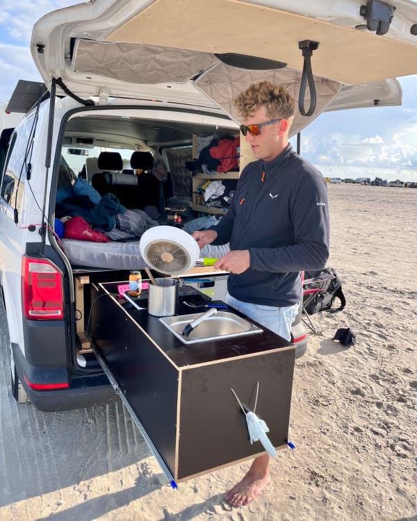 Chris, @van4life__ cooking outdoors at Romo Beach using the custom kitchen setup in his DIY-converted VW T6 camper. The camper's rear hatch is open, showcasing a compact and functional interior with a bed and storage. The sandy beach setting emphasizes the freedom and flexibility of vanlife.