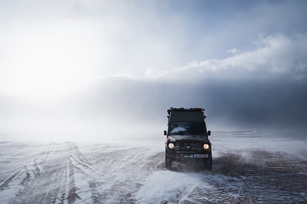 A Puch 230GE 4x4 offroad camper driving through a snowy and foggy landscape in South America, with headlights on, creating a dramatic scene against a misty, cloud-covered background.