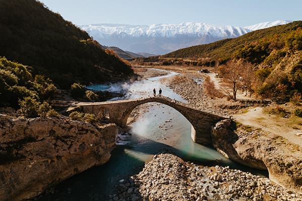 Thomas & Regina, from @edelweiss.on.the.road, are seen crossing a historic stone bridge in Permet, Albania, with snow-capped mountains in the background. This scenic moment highlights their vanlife journey as they explore Albania’s natural beauty and remote landscapes.