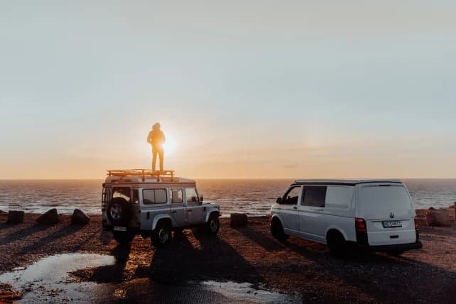 A person standing atop a vintage SUV near the shoreline at sunset, silhouetted against the glowing sun, with another white van parked beside. The serene ocean forms the backdrop with the sun casting a golden hue across the horizon.