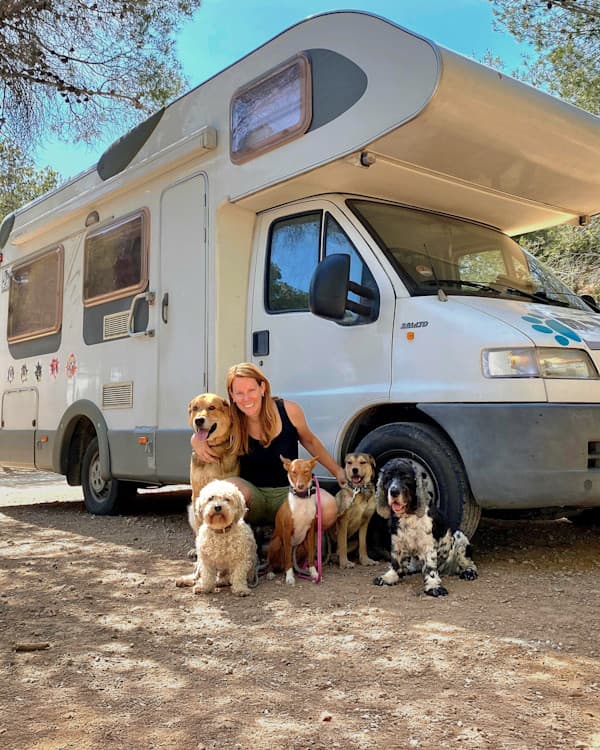 Liz, her five dogs & her campervan "Trevor" - @lizwolting 12/2024 Liz Wolting (@lizwolting), an animal behaviorist and vanlife traveler, poses with her five dogs in front of her motorhome south of Valencia, Spain. The motorhome is parked under the shade of tall trees, and Liz kneels on a dirt ground, smiling alongside her diverse group of dogs, including a golden retriever, a curly-haired small dog, and other mixed breeds. A bright blue sky and natural surroundings in the background.