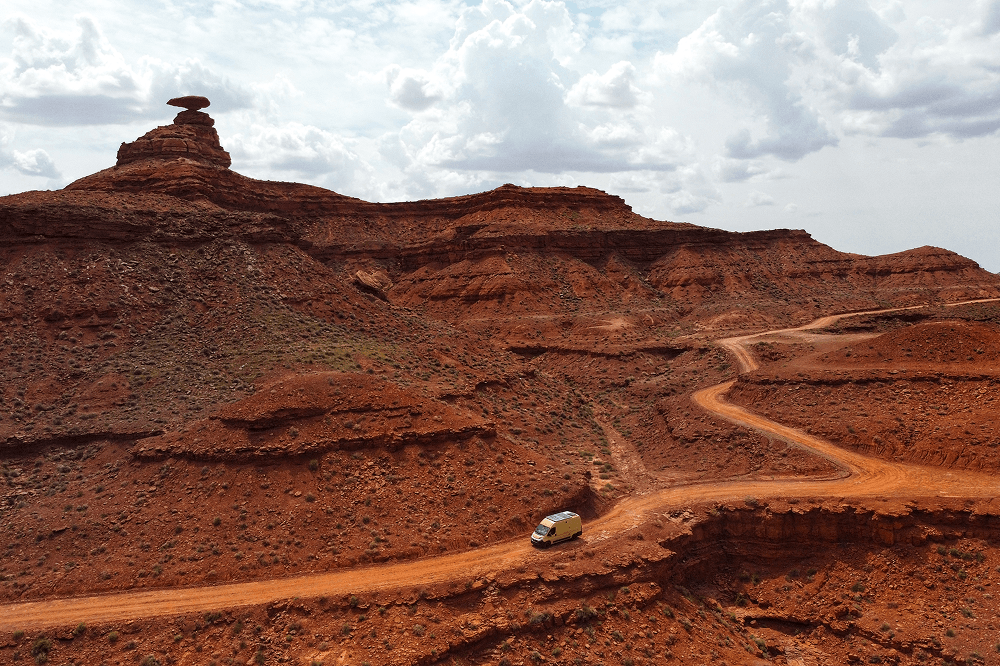 Self converted Fiat Ducato camper van driving along a winding red dirt road through dramatic desert cliffs near Mexican Hat, Utah, captured during a long term vanlife and overlanding journey by @gary_and_sheila. The image shows remote canyon landscapes, rugged off road travel, and life on the road in the American Southwest under a wide cloudy sky.