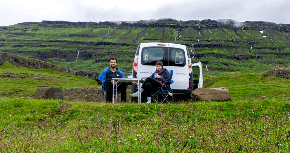 Camping in Iceland - @hayleyandjake 10/2020 A couple enjoying a meal at a portable table with chairs next to a white Renault Kangoo van, parked in the Icelandic countryside with cascading waterfalls in the distance, lush green hills, and overcast skies.