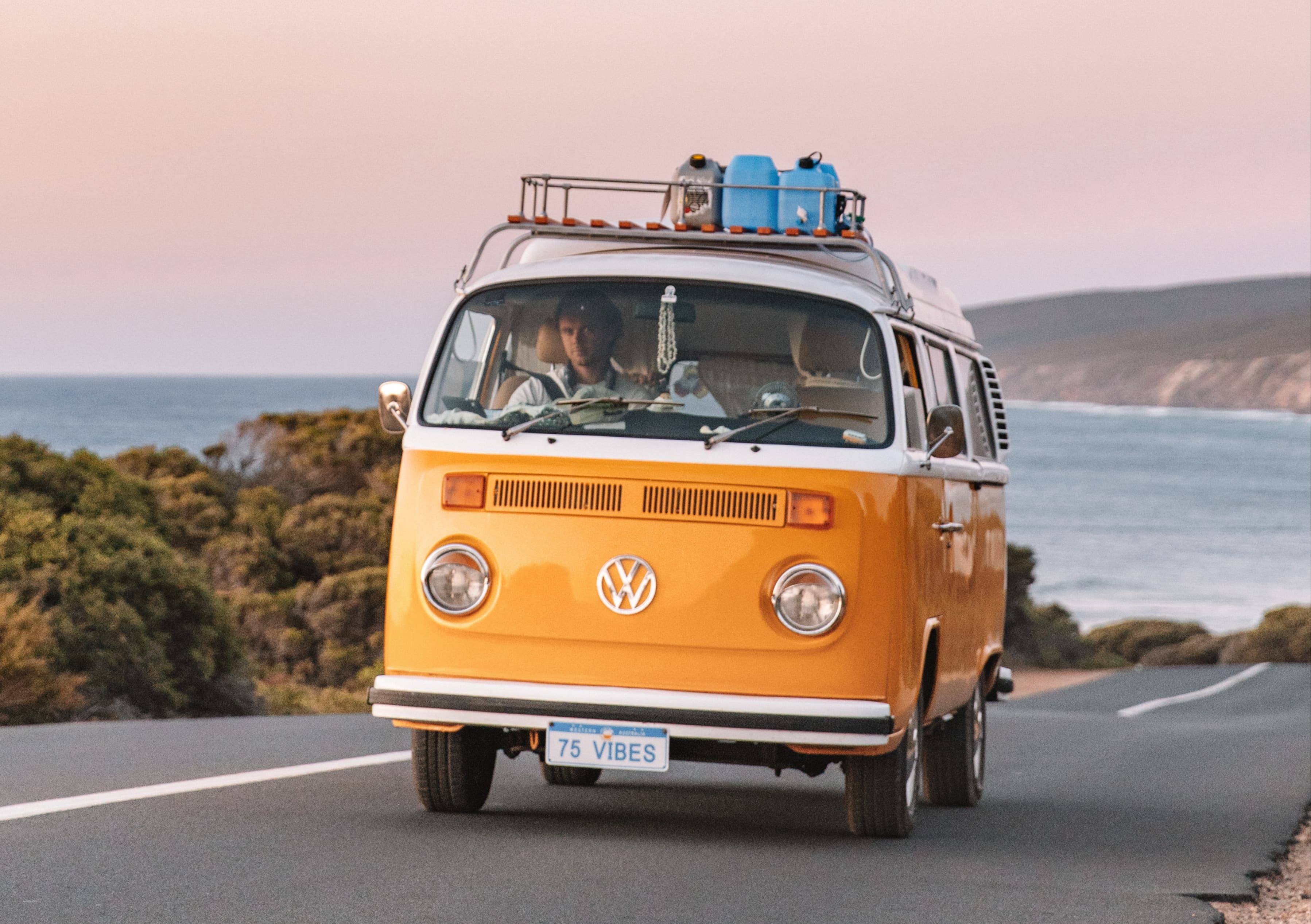 Going with the flow - @75vibes_ 09/2020 A yellow Volkswagen van with the license plate '75 VIBES' drives along a coastal road. Surfboards are visible on the roof rack, hinting at an adventurous beach outing. Inside, a person is focused on the road ahead, while the backdrop features a calm sea and a clear sky at twilight, conveying a sense of freedom and the spirit of a road trip along the picturesque coastline. @75vibes_ 09/2020 - Vanlifezone (Issue 1)